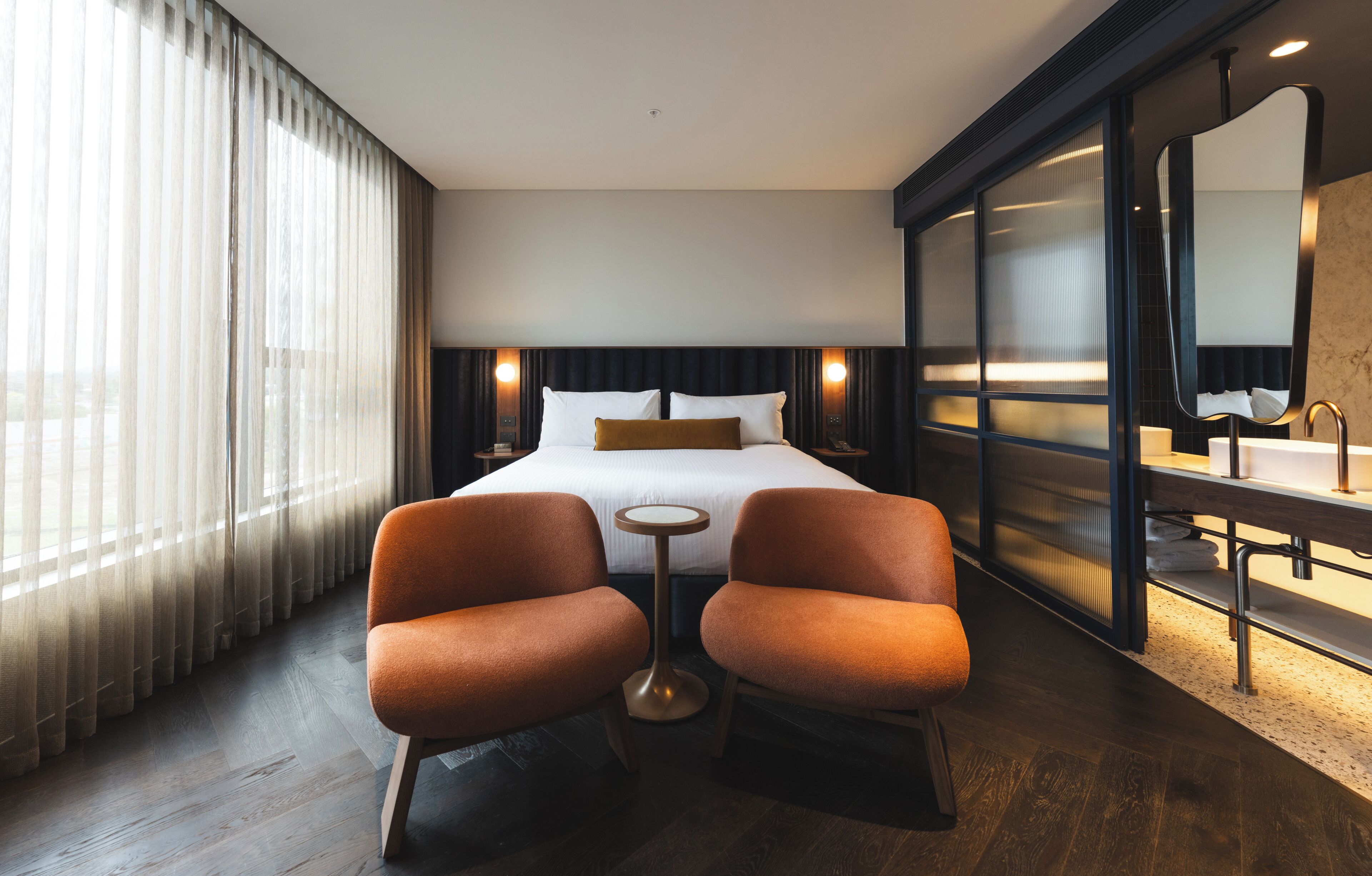A hotel room with a bed, two armchairs, a small table, a large mirror, and a bathroom with a sink and mirror.
