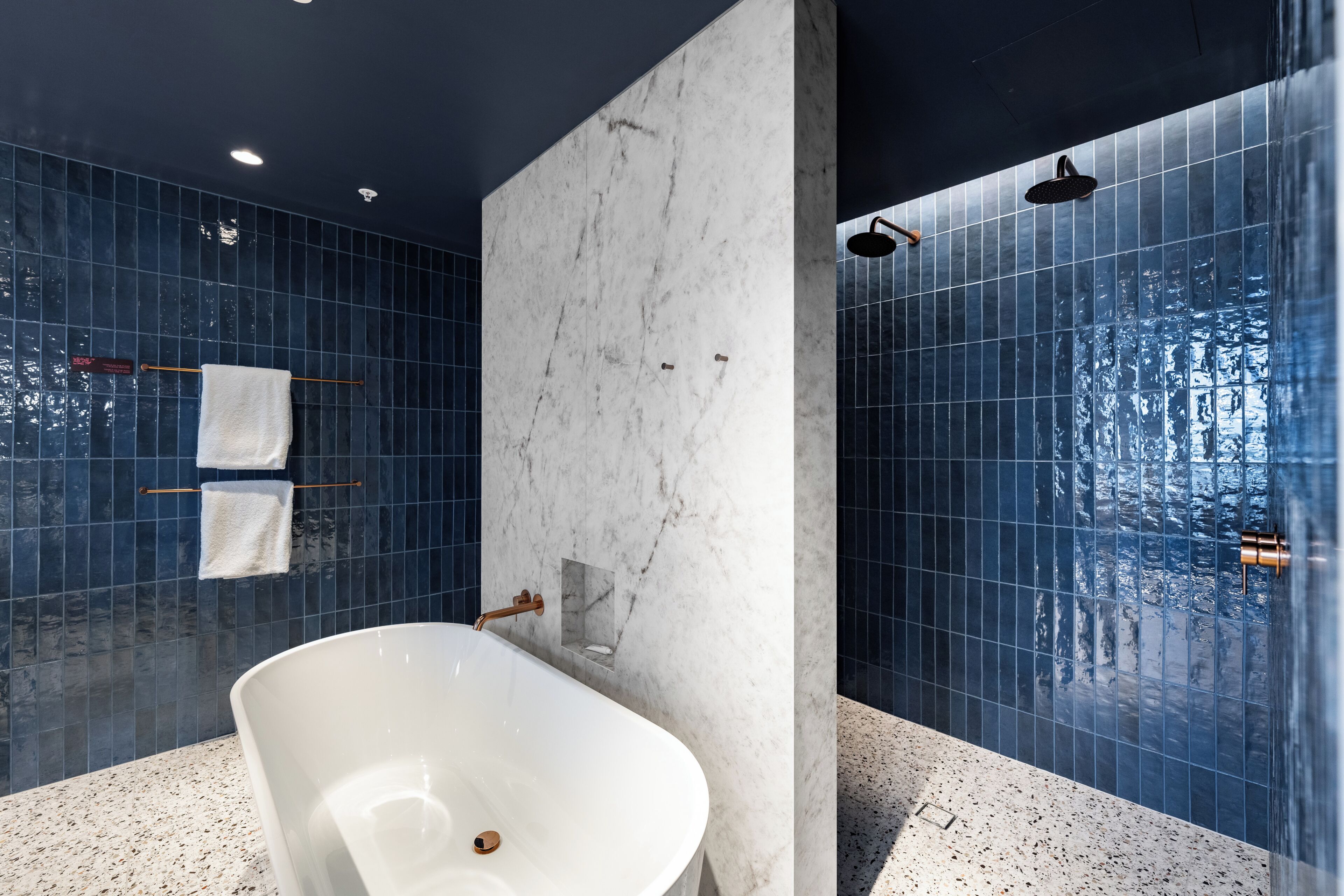 A modern bathroom with a freestanding bathtub, dark blue tiled walls, and a marble accent wall.