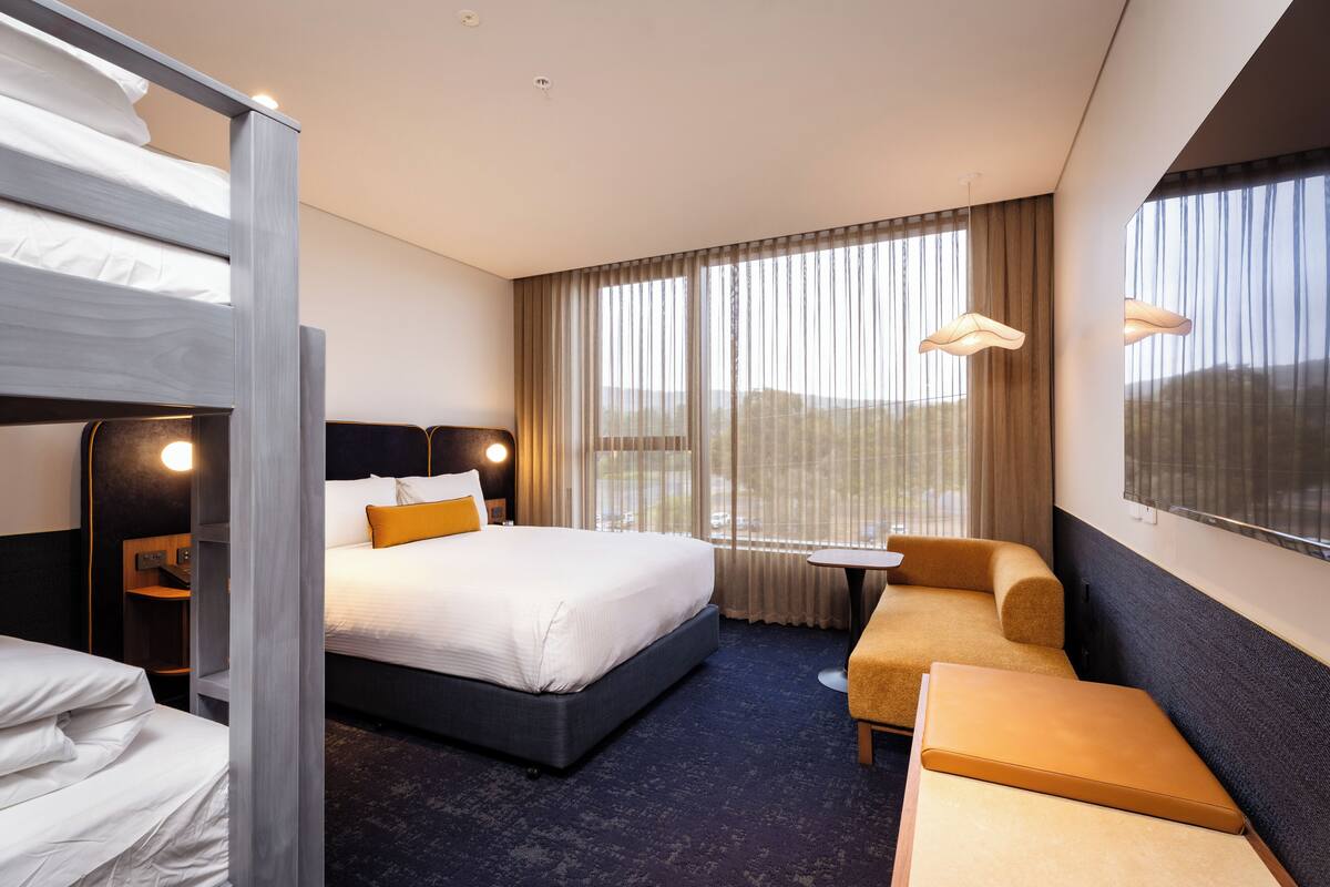 A hotel room with a bunk bed, a single bed, a sofa, a small table, and large windows with curtains.