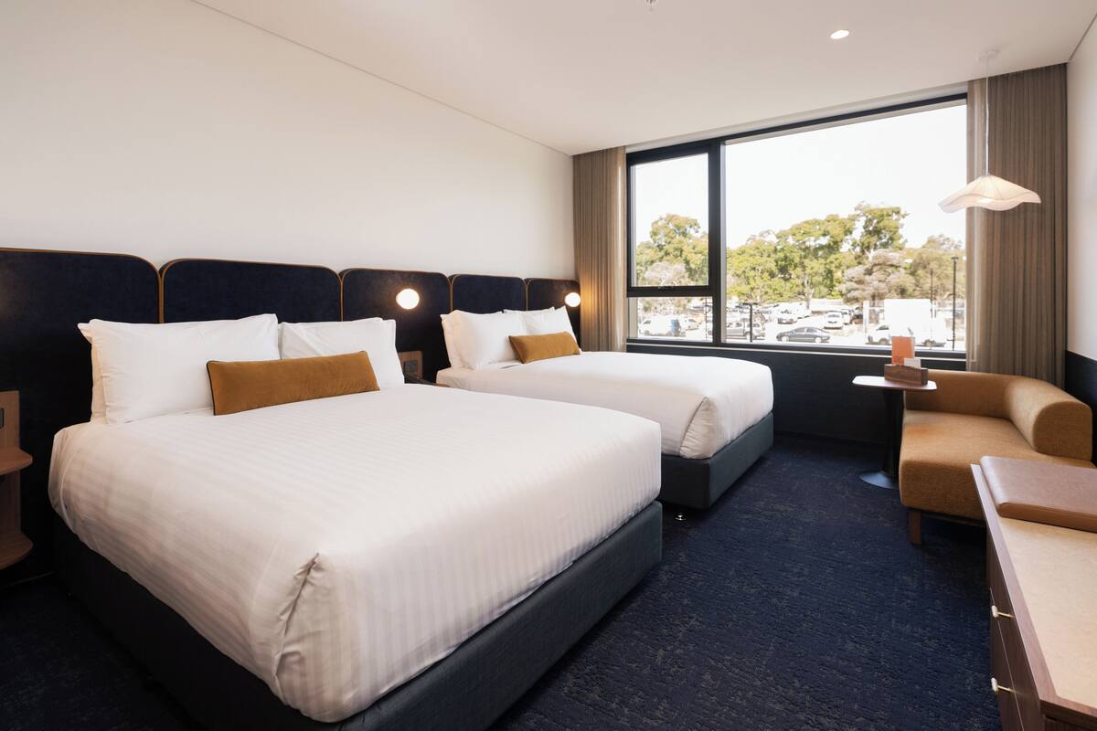 A hotel room with two beds, a sofa, a small table, and a window with a view of trees and parked cars.