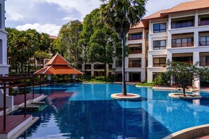 A swimming pool with a wooden deck, surrounded by palm trees and a multi-story building with balconies.