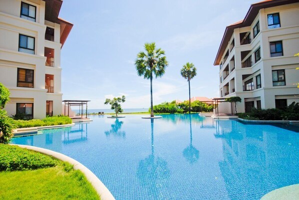 A swimming pool surrounded by modern buildings with balconies, palm trees, and a clear sky.