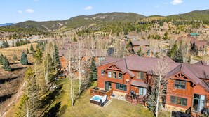 Une grande maison en bois à plusieurs étages, dotée d’un jacuzzi dans le jardin, entourée d’une zone boisée et avec des collines en arrière-plan.