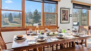 Un coin repas avec une longue table en bois dressée pour le repas, de grandes fenêtres offrant une vue sur un paysage montagneux et une œuvre d’art encadrée accrochée au mur.