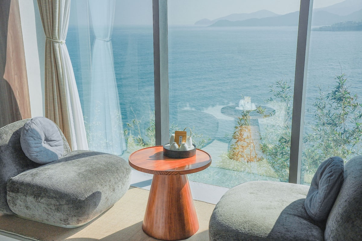 Una habitación moderna con un gran ventanal con vistas a una masa de agua, una mesa redonda de madera y dos sillas grises tapizadas.
