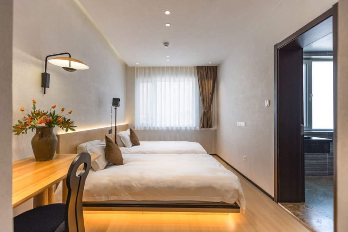 Habitación de hotel con dos camas, un escritorio, una silla, un florero con flores y un ventanal con cortinas.