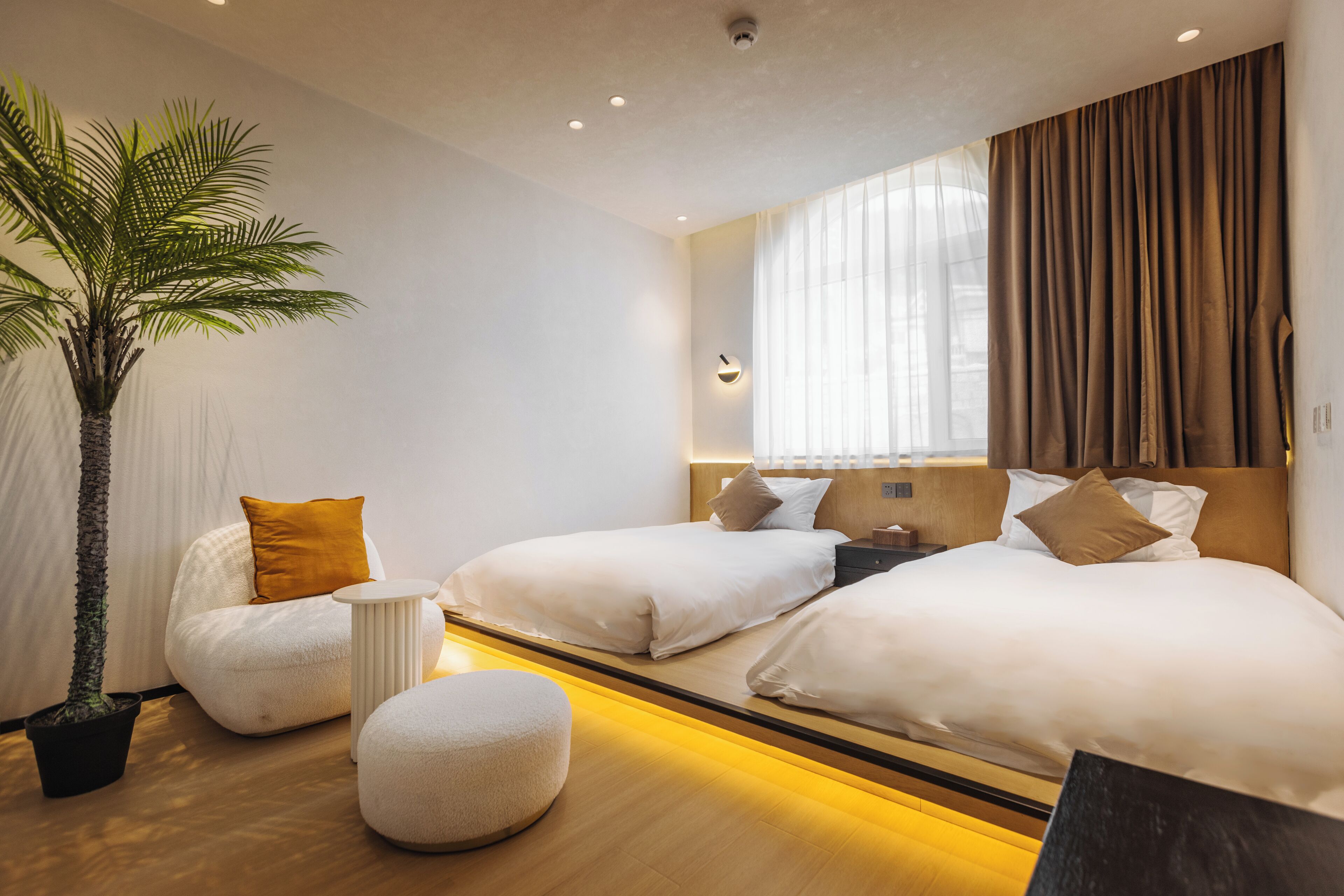 A modern hotel room with two beds, a large window with curtains, a potted palm tree, and a small round ottoman.