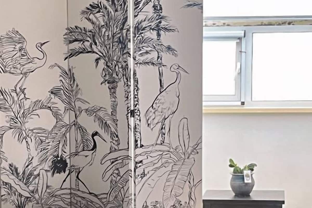 Un dormitorio con dos camas, un mural grande de aves y plantas tropicales, una mesita con una planta, y una ventana con luz natural.