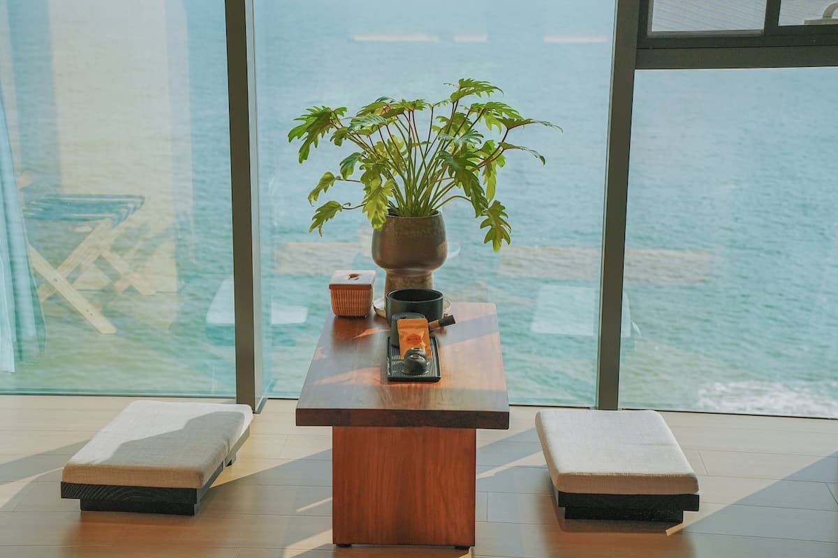Una mesa de madera con una planta en maceta, una tetera y una taza en una habitación moderna con grandes ventanales con vistas al mar.