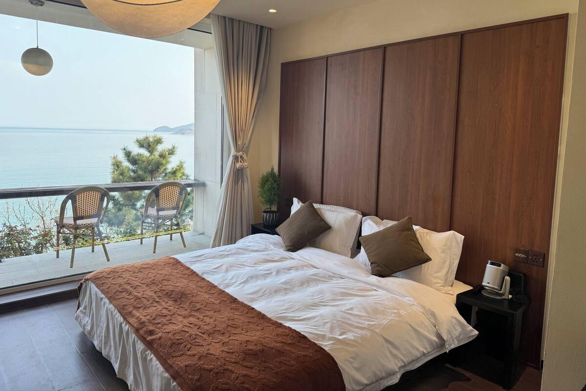 Un dormitorio con una cama grande, una mesita de noche, una lámpara y vista al mar.
