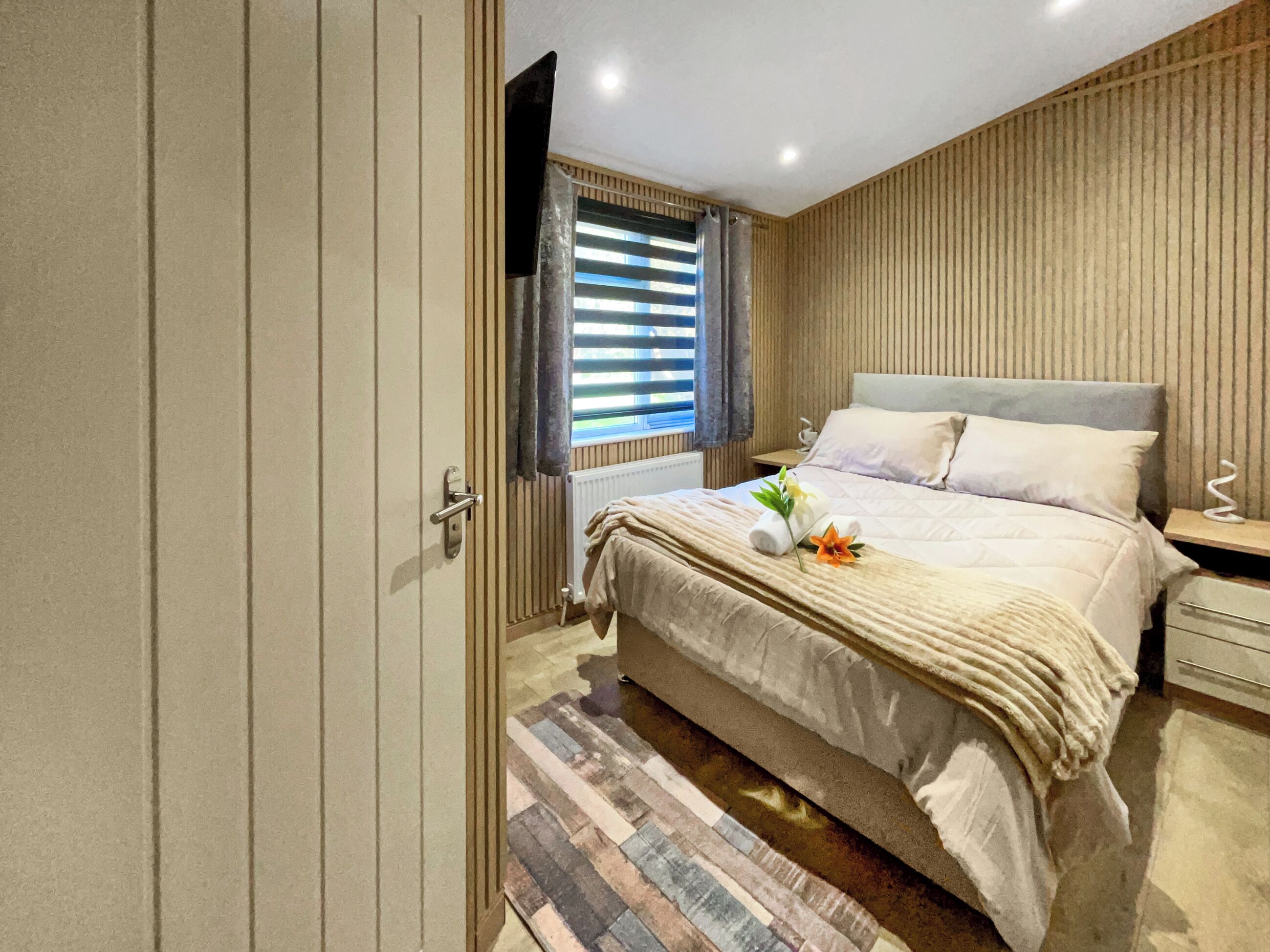 Quarto com cama, mesa de cabeceira, televisão, janela com persianas e porta.