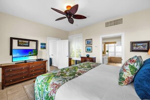 Une chambre à coucher comprenant un lit, un téléviseur, un ventilateur de plafond et une porte donnant sur une salle de bain.