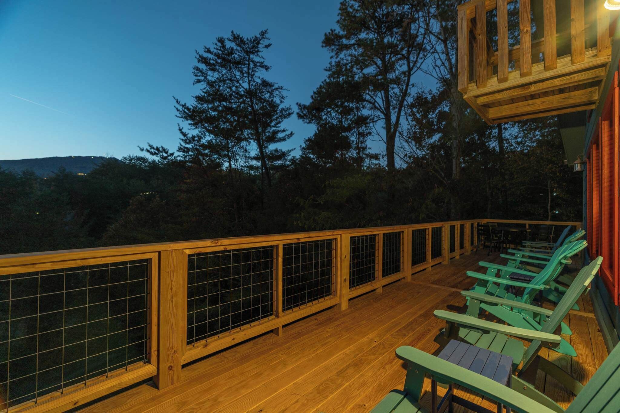 A wooden deck with a railing, green Adirondack chairs, and a view of trees and hills.