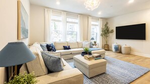 En moderne stue med sofa, skammel og sofabord med bøger. Der er et stort vindue med gardiner, en gulvlampe og en potteplante.