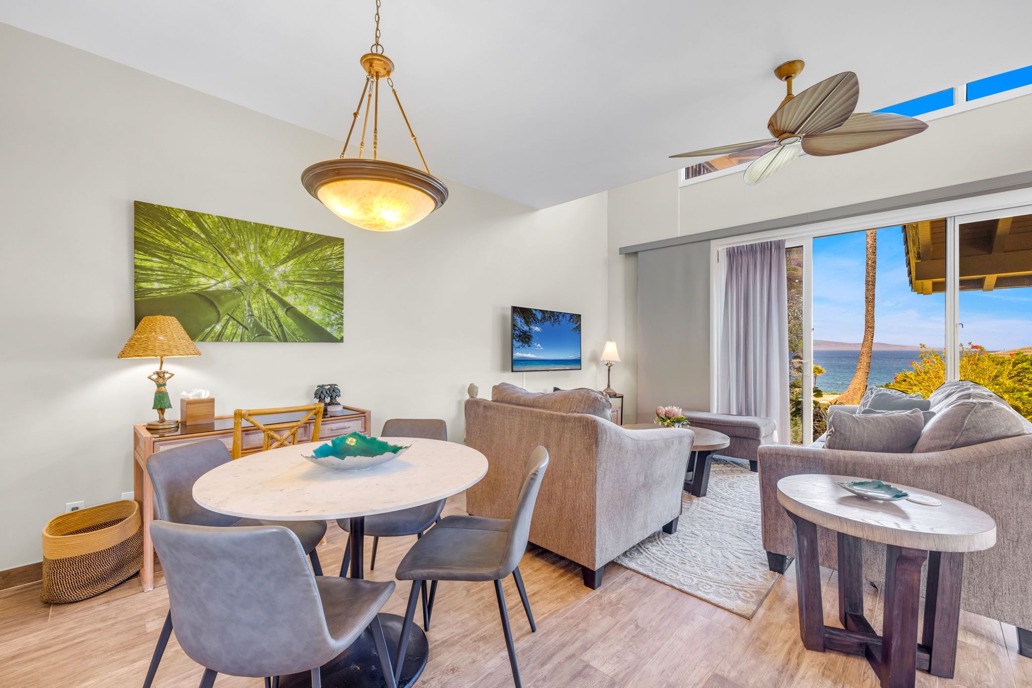 A modern living room with a dining area, a ceiling fan, and a large window with a view of the ocean.