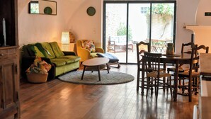 Una sala de estar acogedora con un sofá verde, un sillón amarillo y una mesa de centro redonda. Hay una zona de comedor con sillas de madera y una mesa. Un amplio ventanal corredero da a un patio exterior.