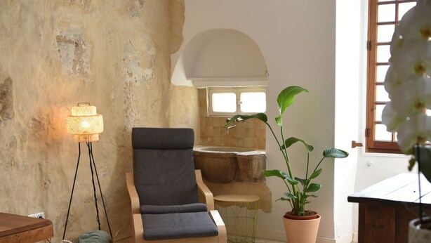 Una habitación acogedora con una pared rústica, un sillón con cojines, una lámpara de pie, una planta en maceta y una mesa de madera.