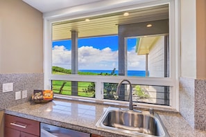 A kitchen with a stainless steel sink, a window overlooking a grassy area, and a bag of Hawaiian coffee on the counter. - Kapalua Bay | Ocean View 2 BR, Sleeps 6 | Car Incl. w/ 6+ Nights | KBV ML-1984 by KBM (Lahaina)