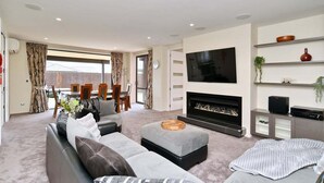 Una sala de estar moderna con un conjunto de sofás, una chimenea, un televisor montado en la pared y un comedor visible a través de la ventana.