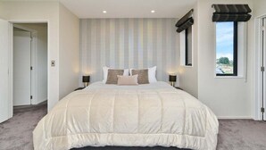 Un dormitorio con una cama grande, dos mesitas de noche, un ventanal con vistas y una puerta que conduce a otra habitación.