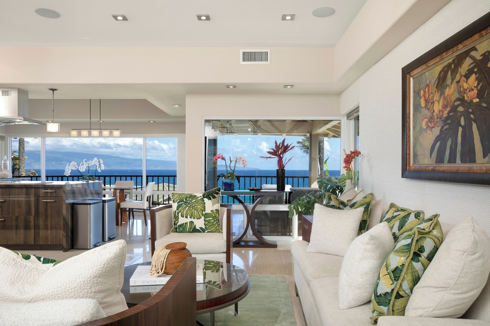 A modern living room with a view of the ocean, a dining area with a glass table, and a kitchen with stainless steel appliances.