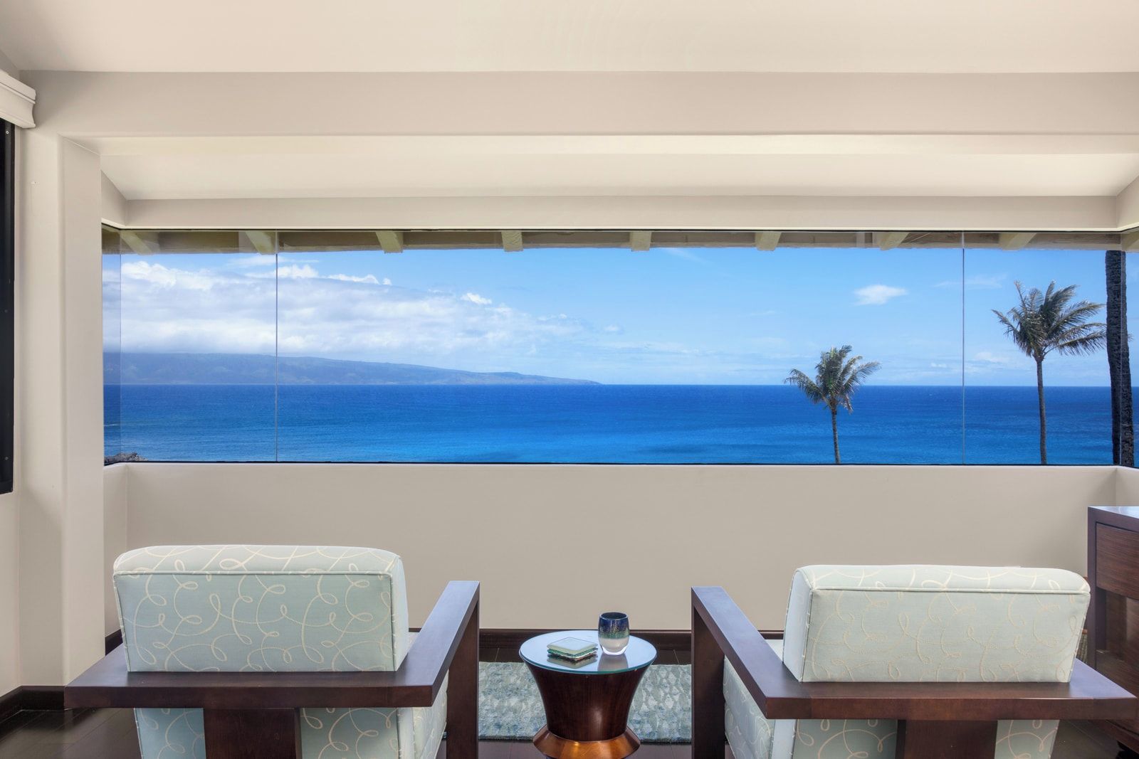 A room with a large window overlooking the ocean, two chairs, a small table, and a view of palm trees.