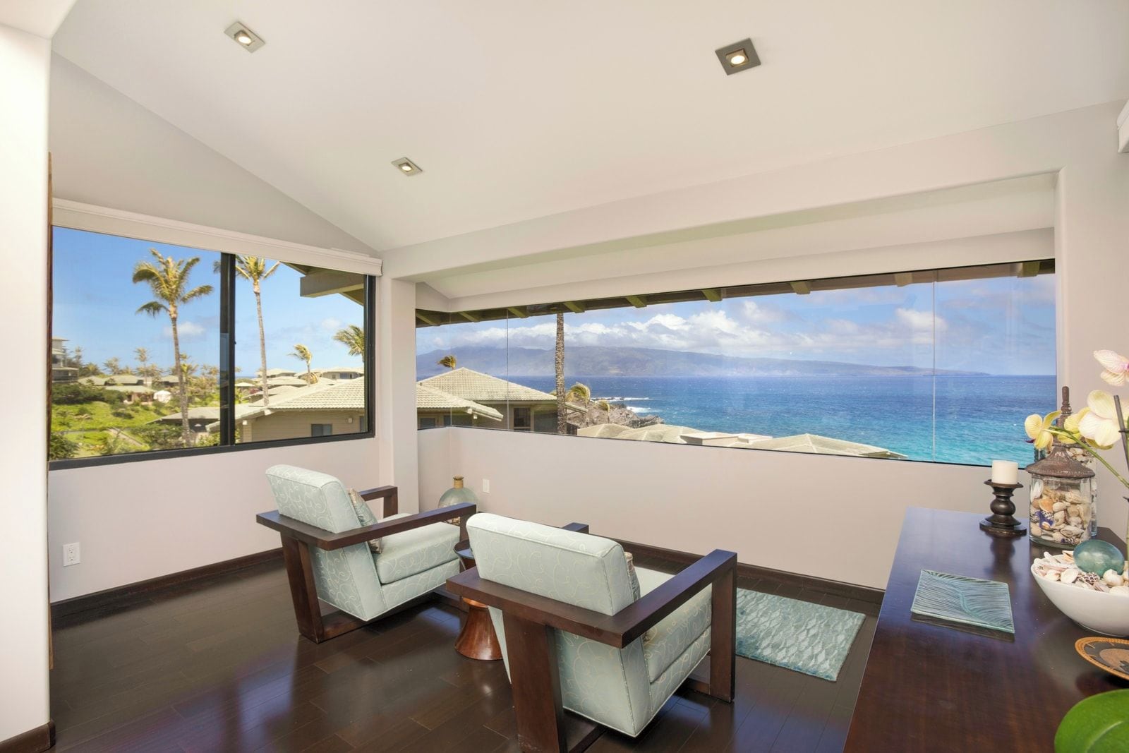 Ein Zimmer mit einem großen Fenster, das Blick auf das Meer und Palmen bietet. Zwei Sessel und ein kleiner Tisch mit Dekorationen sind vorhanden.
