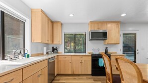 A modern kitchen with wooden cabinets, a stainless steel microwave, and a dining area with a wooden table and chairs. - Jed Smith Cabin with Tennis/Pickle ball courts and HOT TUB! (Crescent City)