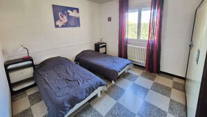 Una stanza con due letti, un termosifone, una finestra con tende, una scrivania con una lampada e un quadro appeso al muro.
