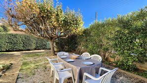 Zona pranzo all'aperto con tavolo apparecchiato per quattro, circondata da sedie bianche, in un giardino con pavimentazione in ghiaia e diverse piante.