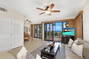 A spacious living room with a large TV, a ceiling fan, and a view of the ocean through the sliding glass doors. - Kapalua Bay | Ocean View 3 BR, Sleeps 10 | Car Incl. w/ 6+ Nights | KBV ML-1987 by KBM (Lahaina)