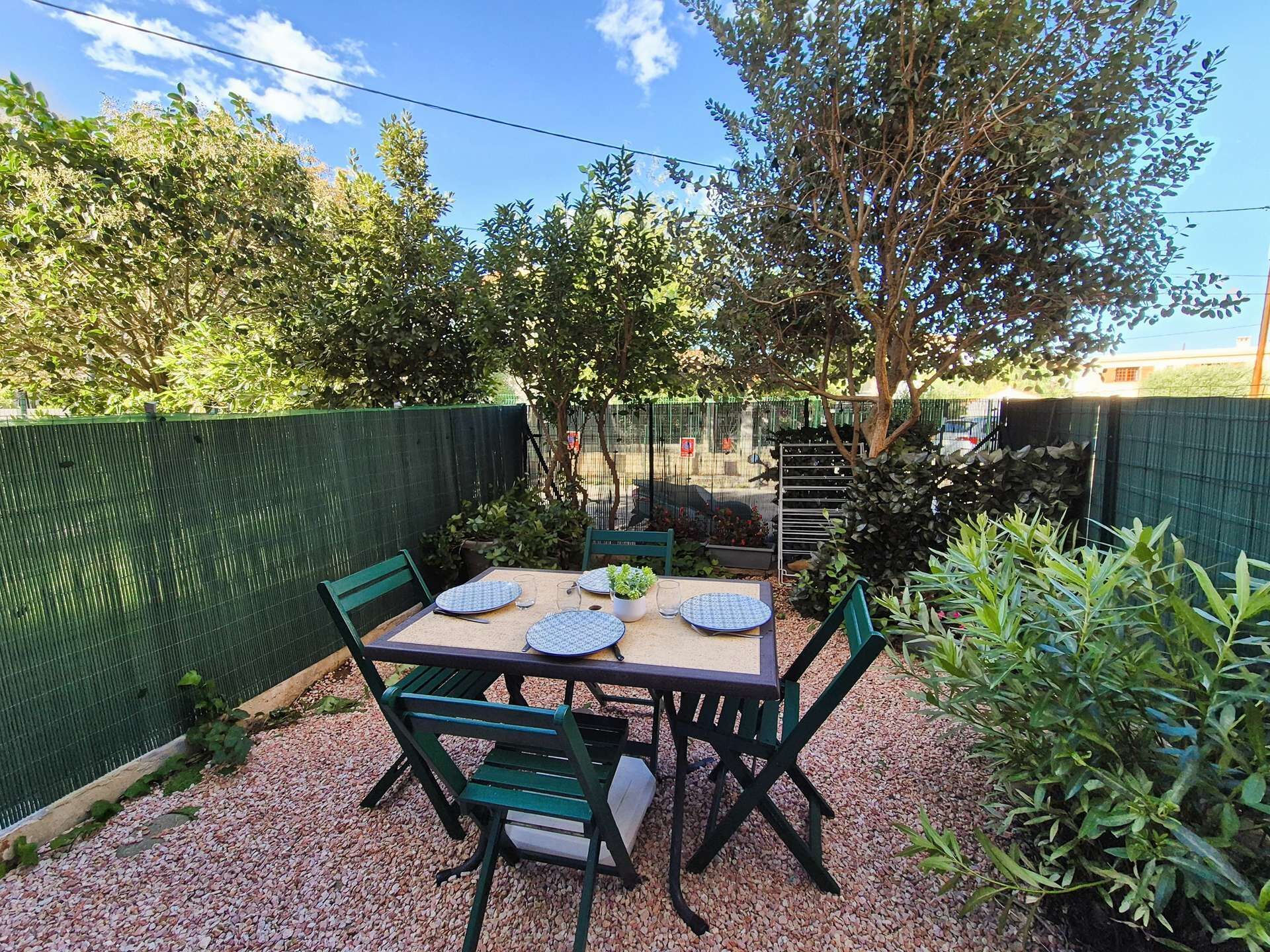 An outdoor patio with a table and chairs, surrounded by greenery and a green fence.