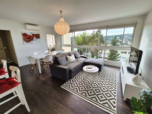 A modern living room with a sofa, coffee table, and dining area. There is a large window offering a view of the city. - 2-bedroom apartment, sea view, A/C, WiFi, pool (ST MANDRIER SUR MER)