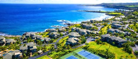 An aerial view of a coastal residential area with houses, a tennis court, and a view of the ocean.