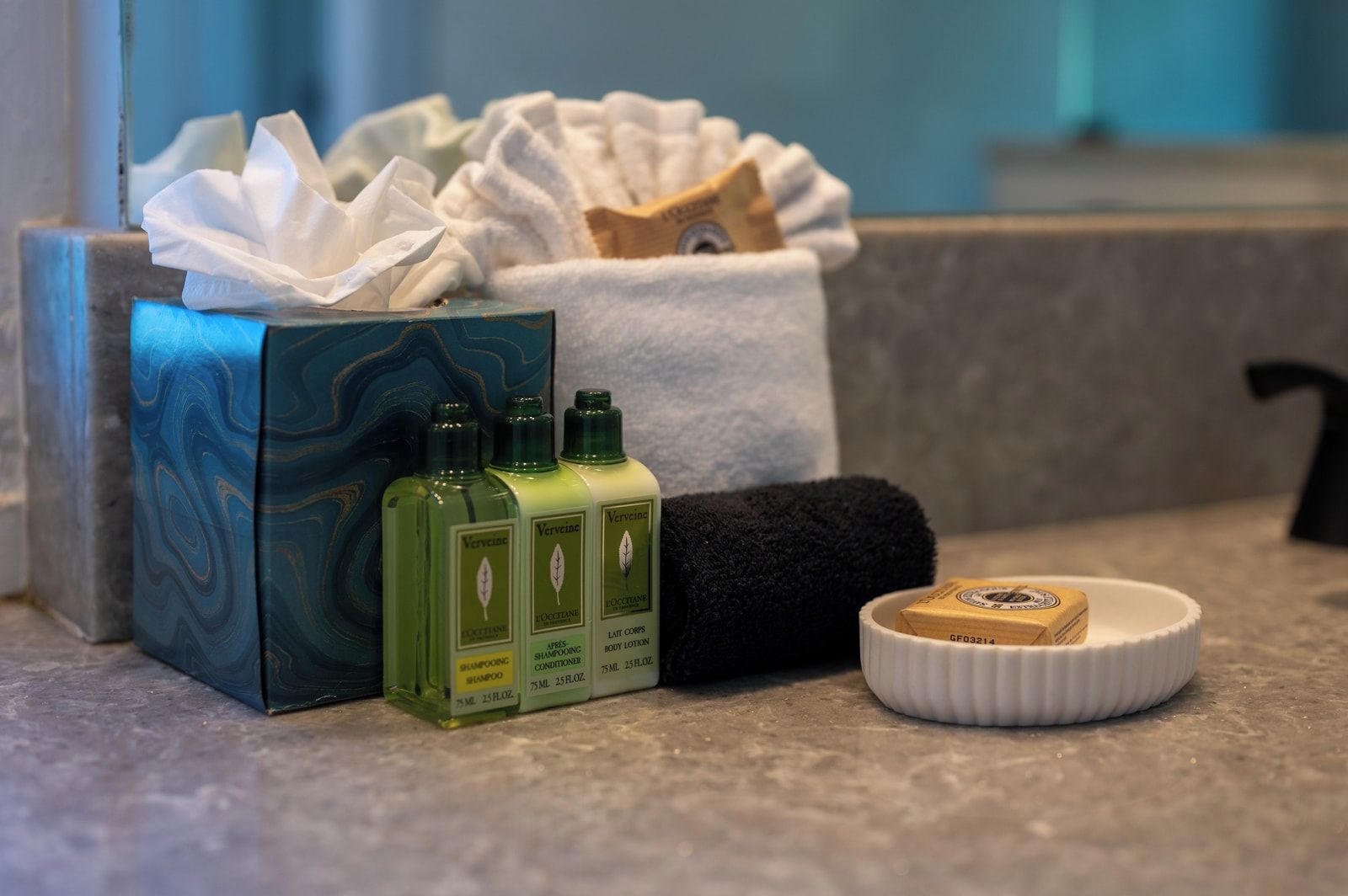 A bathroom counter with a box of toiletries, three bottles of shampoo, a black towel, and a soap dish.