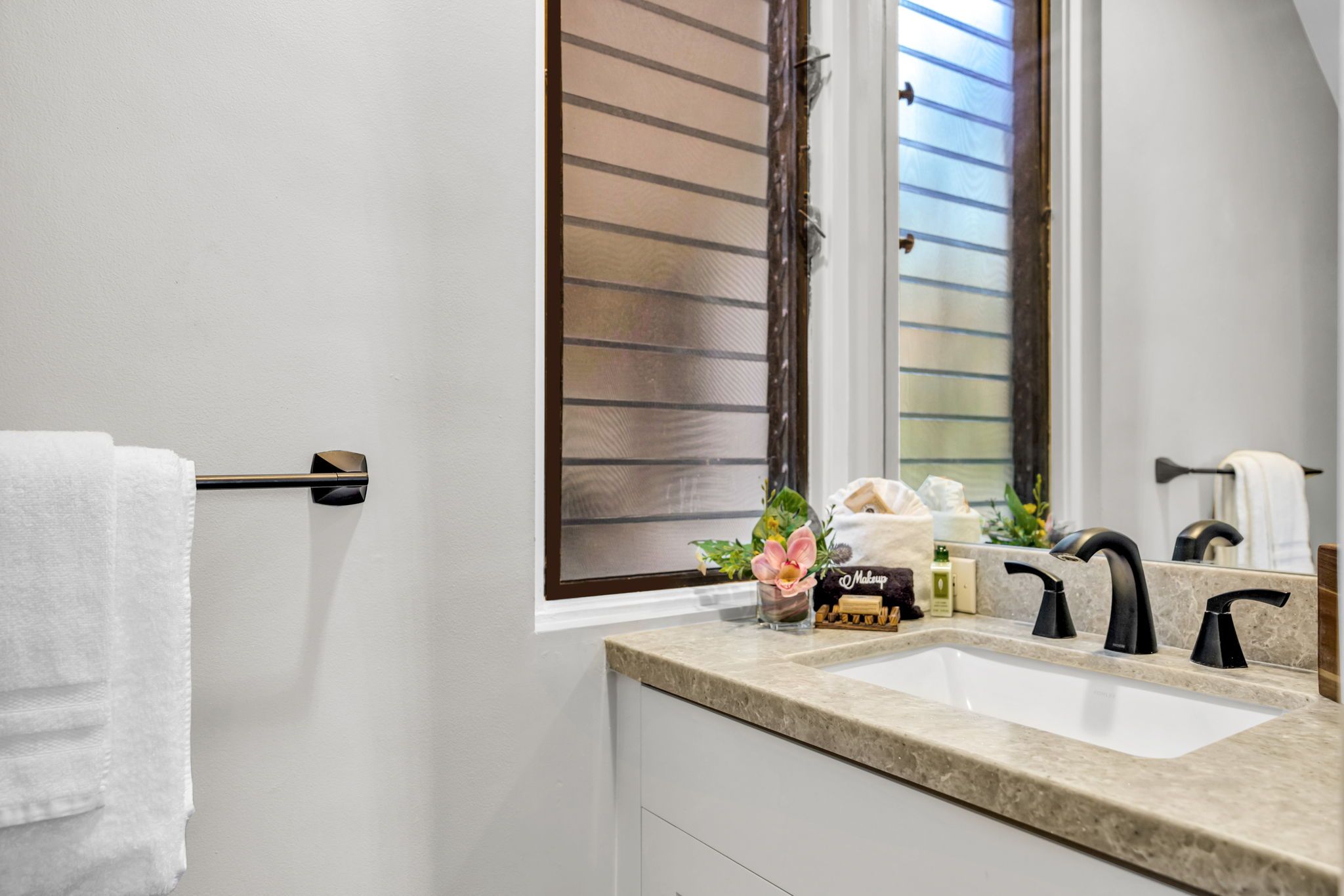 A bathroom with a white sink, black faucets, a towel rack, a window with blinds, and a small plant on the counter.