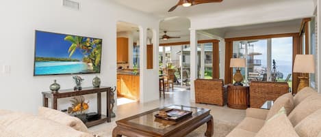 A spacious living room with a large sofa, a coffee table, and a television mounted on the wall. There is a dining area visible through an open doorway, and a balcony with a view of the ocean.