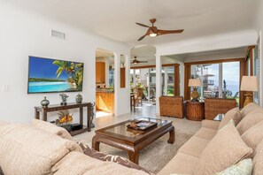 A spacious living room with a large sofa, a coffee table, and a television mounted on the wall. There is a dining area visible through an open doorway, and a balcony with a view of the ocean. - Kapalua Bay | Ocean View 2 BR, Sleeps 8 | Car Incl. w/ 6+ Nights | KBV ML-2008 by KBM (Lahaina)