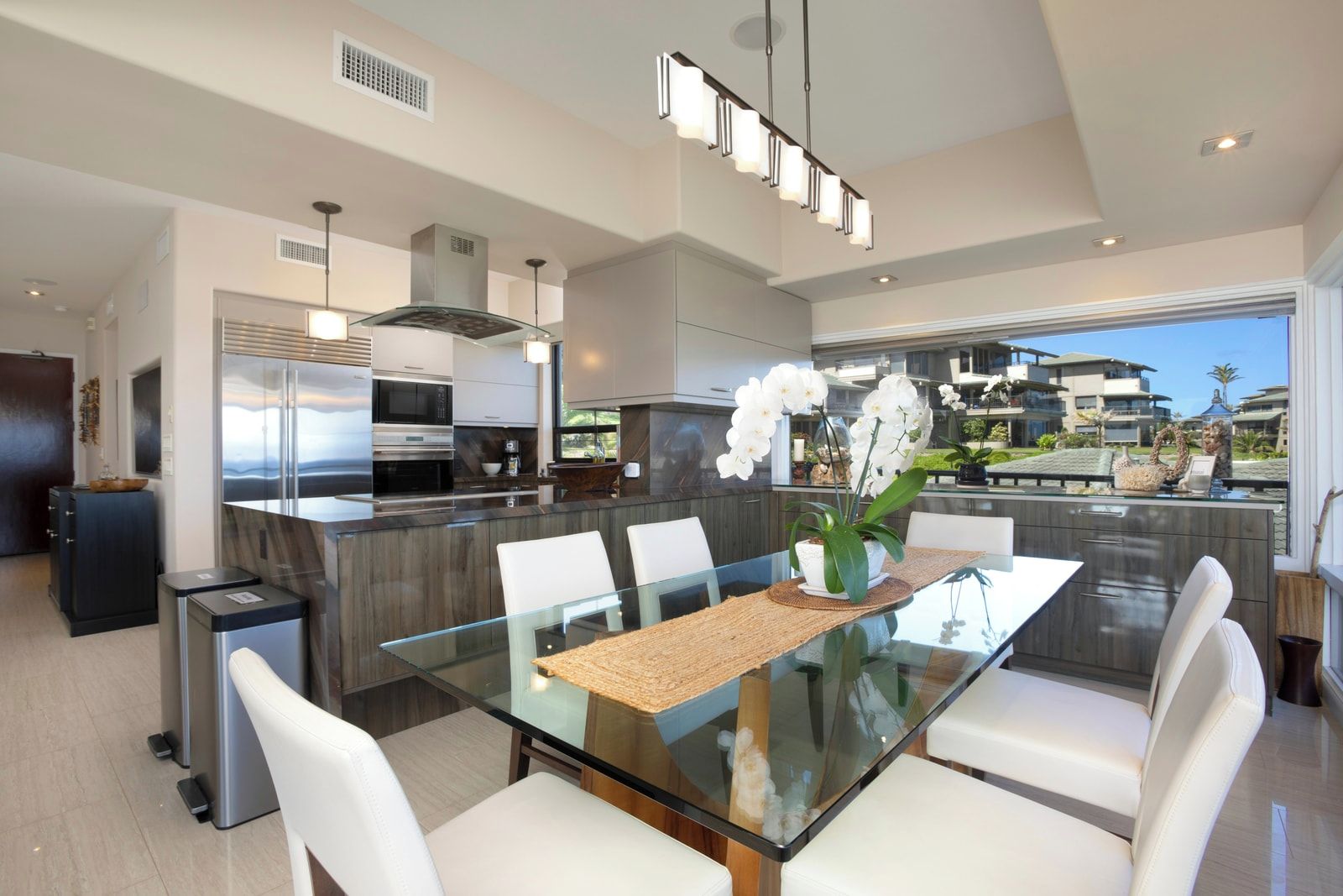 A modern kitchen with a glass dining table, white chairs, and a view of a residential area.