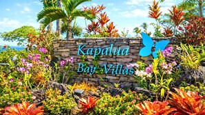 A stone sign with "Kapalua Bay Villas" written on it, surrounded by tropical plants and palm trees. - Kapalua Bay | Ocean View 2 BR, Sleeps 8 | Car Incl. w/ 6+ Nights | KBV ML-1996 by KBM (Lahaina)