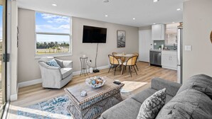 Una sala moderna con un sofá gris, un sillón blanco y una mesa de centro con un cuenco. Hay un comedor con una mesa y sillas, y una cocina con armarios blancos y un refrigerador.