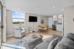 Ein modernes Wohnzimmer mit grauem Sofa, einem weißen Sessel und einem Couchtisch mit einer Schale. Es gibt einen Essbereich mit Tisch und Stühlen sowie eine Küche mit weißen Schränken und einem Kühlschrank.