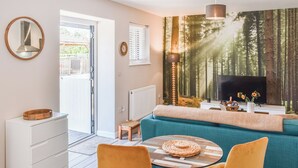 En moderne stue med sofa, to lænestole, et sofabord, et fjernsyn og et stort vægmaleri med skovmotiv.