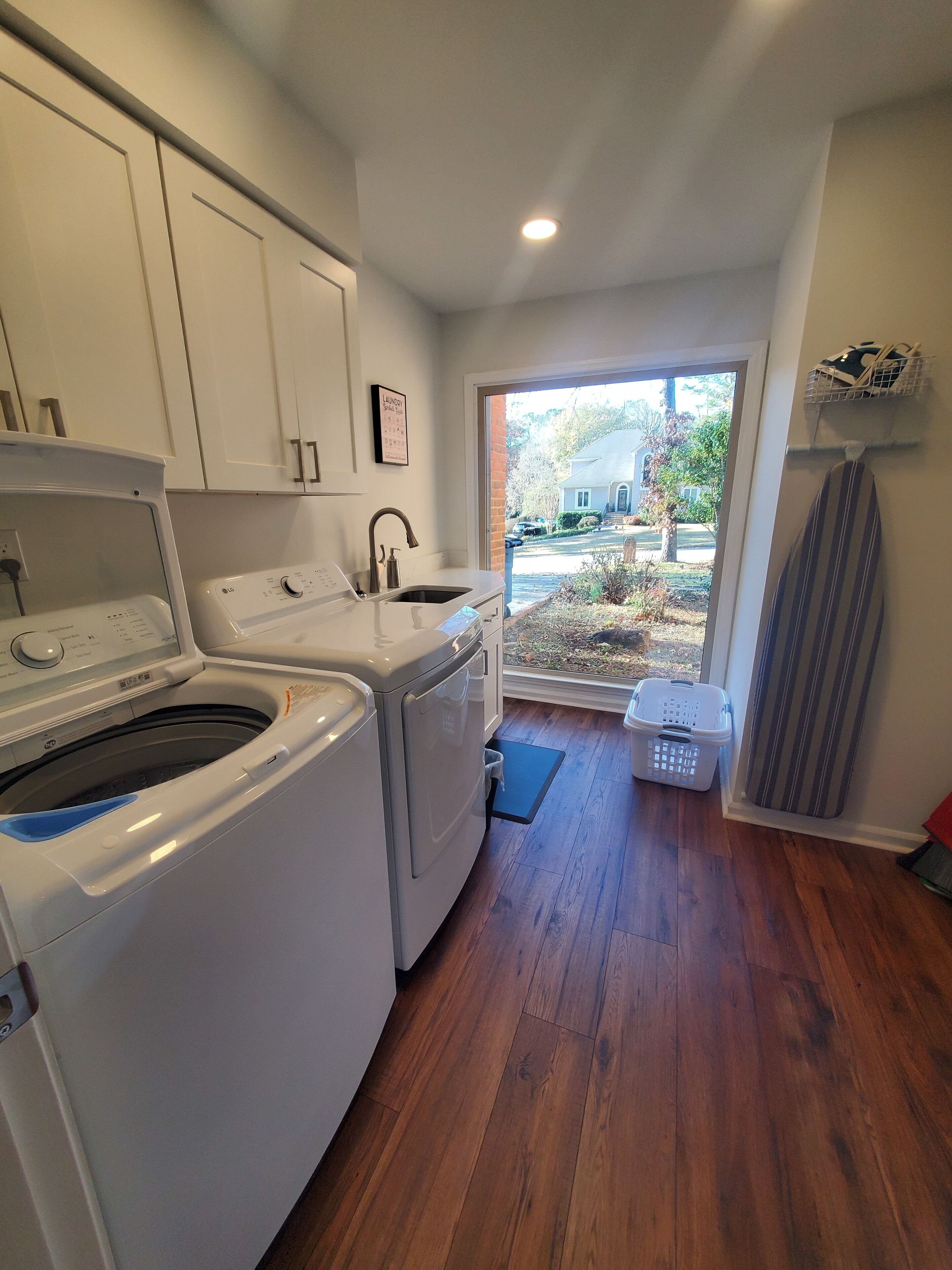 A laundry room with a washing machine, dryer, and sink. There is a window with a view of a house and trees.