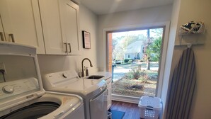 Un lavadero equipado con lavadora, secadora y lavabo. Se ve una ventana con vistas a una casa y árboles.