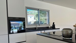 Una cocina moderna con horno integrado, una ventana con vistas a una casa y árboles, encimera negra y un jarrón con flores secas.