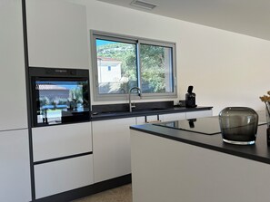 A modern kitchen with a built-in oven, a window with a view of a house and trees, a black countertop, and a vase with dried flowers. - Villa Rosula private pool, heated (Oletta)