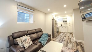 Een moderne woonkamer met een bruine leren bank, een witte salontafel en een afstandsbediening erop. In de achtergrond is een keuken te zien met witte kasten en roestvrijstalen apparatuur.