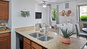 Una cocina con encimeras de granito, lavavajillas, armarios de madera, ventilador de techo, comedor con mesa y sillas, y ventana con vista a la vegetación.