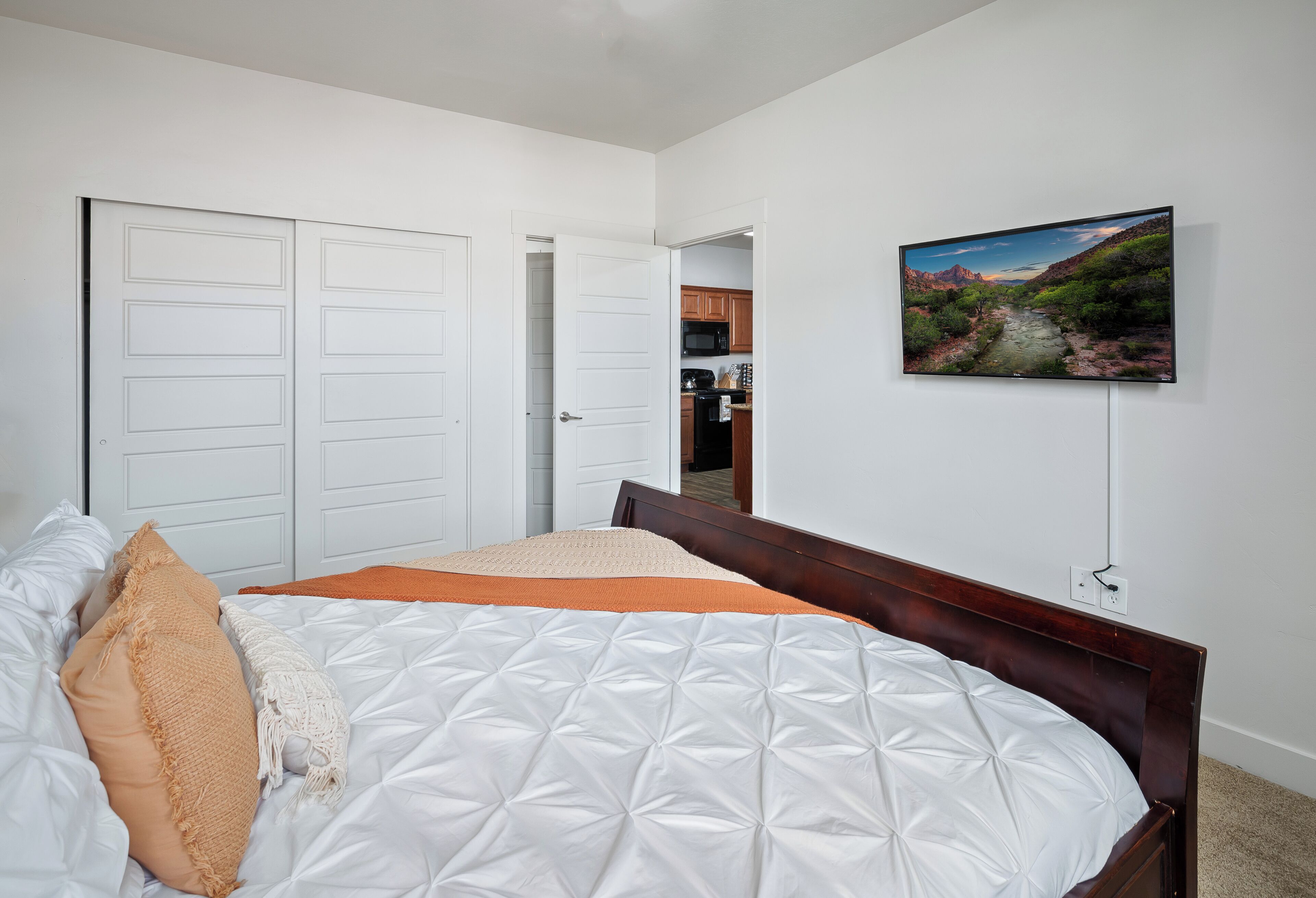 A bedroom with a bed, a TV, and a door leading to a kitchen.
